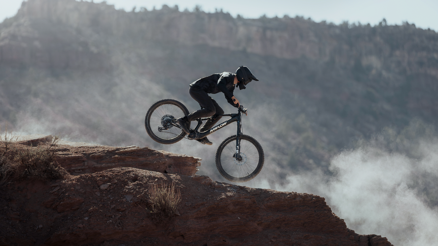 A mountain biker in black riding gear jumps a black mountain bike over a rocky canyon edge, with mist and a rugged desert landscape in the background.